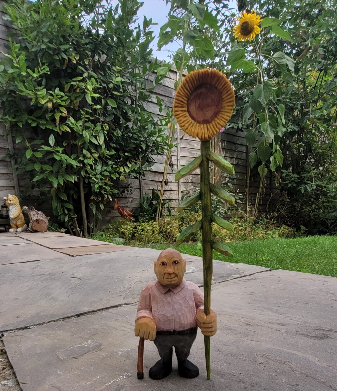 Carved figure holding a tall sunflower, standing in a garden